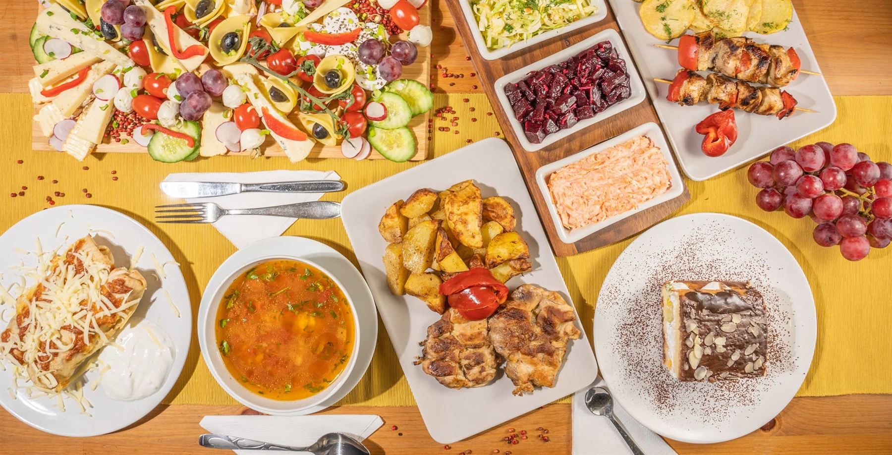 Masă întinsă pe un napron galben: ciorbă, pui la grătar cu cartofi, frigărui, salate de sfeclă, varză și morcov, platou de brânzeturi și prăjitură.